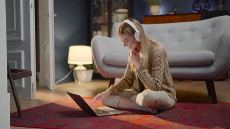 Happy-Young-Woman-With-Wireless-Headphones-Chatting-On-Laptop-Computer-While-Sitting-On-Floor-At-Home