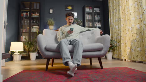 Junger-Mann-Mit-Smartphone,-Während-Er-Zu-Hause-Auf-Einer-Couch-Sitzt-1