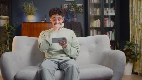Young-Man-Watching-Some-Unexpected-Movie-Scene-On-Smartphone-While-Sitting-On-Couch-At-Home