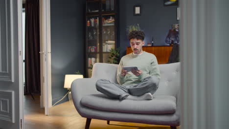 Young-Man-Sitting-On-Sofa-With-Crossed-Legs-And-Watching-Something-On-Smartphone