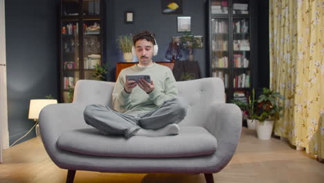 Young-Man-Sitting-On-Sofa-Wearing-Wireless-Headphones-And-Watching-Some-Shocking-Movie-Scene-On-Smartphone