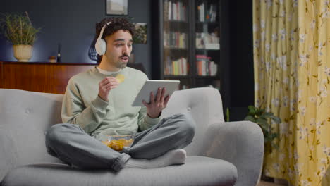 Young-Man-Watching-Unexpected-Movie-Scene-On-Tablet-While-Eating-Chips-And-Sitting-With-Crossed-Legs-On-Couch-At-Home