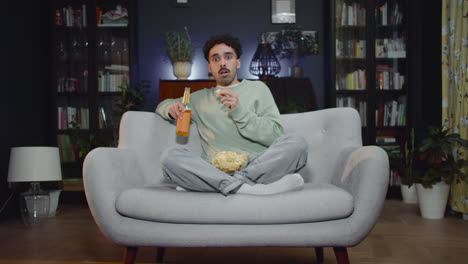 Young-Man-Watching-Unexpected-Movie-Scene-On-Tv-While-Eating-Chips-And-Drink-Soda-Sitting-With-Crossed-Legs-On-Couch-At-Home