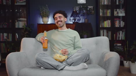 Young-Man-Watching-Comic-Movie-On-Tv-While-Eating-Chips-And-Drink-Soda-Sitting-With-Crossed-Legs-On-Couch-At-Home