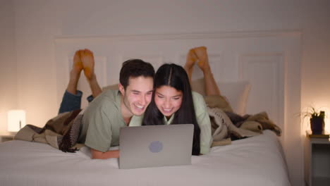Couple-Watching-Comic-Movie-On-Laptop-Lying-In-Bed-At-Home