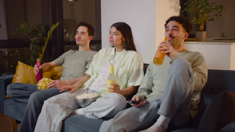 Side-View-Of-Three-Friends-Watching-Interesting-Movie-On-Television-Sitting-On-Couch,-Eating-Popcorn-And-Chips-And-Drinking-Soda