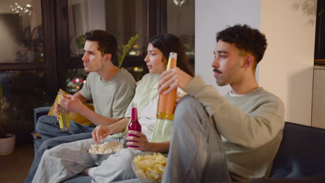 Side-View-Of-Three-Friends-Watching-Interesting-Movie-On-Television-Sitting-On-Couch,-Eating-Popcorn-And-Chips-And-Drinking-Soda-1