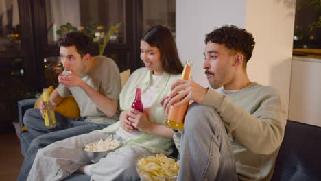 Seitenansicht-Von-Drei-Freunden,-Die-Interessante-Filme-Im-Fernsehen-Sehen,-Auf-Der-Couch-Sitzen,-Popcorn-Und-Chips-Essen-Und-Limonade-Trinken-2