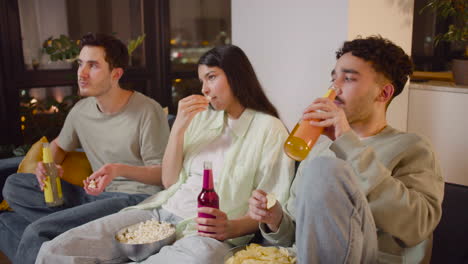 Three-Friends-Watching-Interesting-Movie-On-Television-Sitting-On-Couch,-Eating-Popcorn-And-Chips-And-Drinking-Soda-2