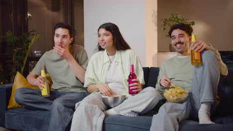 Three-Friends-Watching-Interesting-Movie-On-Television-Sitting-On-Couch,-Eating-Popcorn-And-Chips-And-Drinking-Soda-3