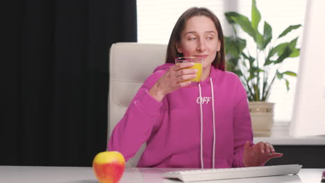 Young-Woman-Drinking-An-Healthy-Orange-Juice-While-Using-Desktop-Computer-At-Office