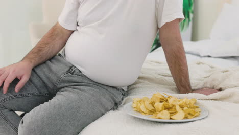 Unrecognizable-Boy-Touching-His-Belly-Fat-Sitting-On-The-Bed-Near-Potato-Chips