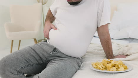 Boy-Touching-His-Belly-Fat-Sitting-On-The-Bed-Near-Potato-Chips