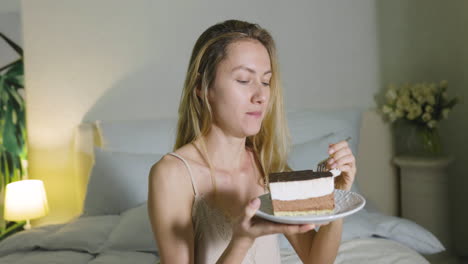 Blonde-Girl-Holding-And-Eating-A-Piece-Of-Cake-Sitting-On-Bed