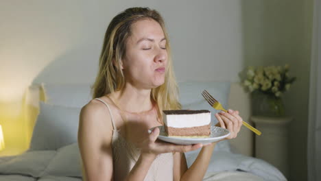 Blonde-Girl-Holding-And-Eating-A-Piece-Of-Cake-Sitting-On-Bed-1