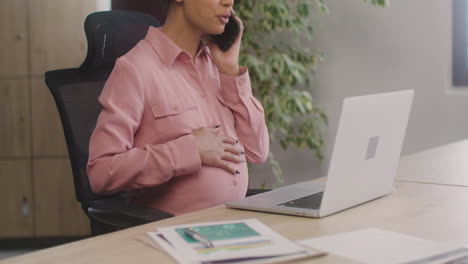 Nahaufnahme-Einer-Schwangeren-Frau,-Die-Mit-Ihrem-Handy-Spricht-Und-Ihren-Bauch-Streichelt,-Während-Sie-Im-Büro-Arbeitet