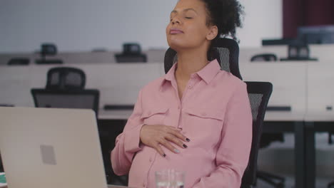 Happy-Pregnant-Woman-Caressing-Her-Belly-While-Sitting-At-Desk-In-The-Office
