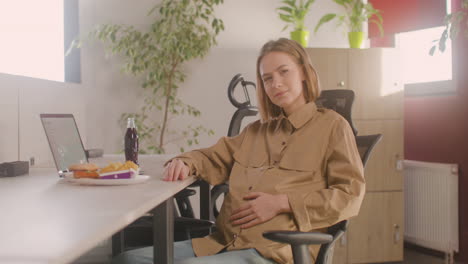 Working-Pregnant-Woman-Sitting-At-Desk-And-Smiling-At-Camera-During-Office-Lunch-Break
