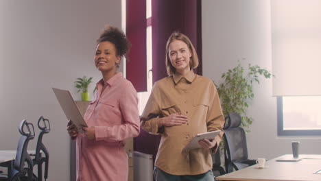 Two-Happy-Pregnant-Woman-Posing-At-Camera-While-Touching-Her-Bellies-And-Holding-Documents-In-The-Office