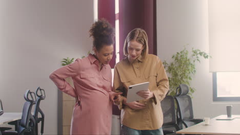 Two-Pregnant-Woman-Looking-At-Tablet-And-Talking-In-The-Office