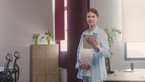 Schwangere-Frau-Posiert-Vor-Der-Kamera,-Während-Sie-Ihren-Bauch-Berührt-Und-Im-Büro-Ein-Tablet-Hält