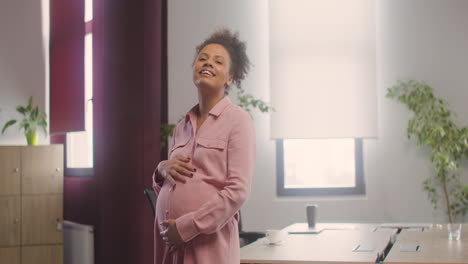 Mujer-Feliz-Embarazada-Posando-En-La-Cámara-Mientras-Toca-Su-Vientre-En-La-Oficina