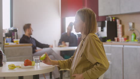 Pregnant-Woman-Sitting-At-Table-Eating-Fruit-And-Talking-With-Female-Coworker-In-Canteen