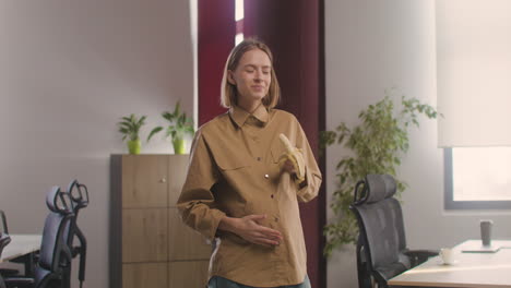 Pregnant-Happy-Woman-Standing-Eating-A-Banana-And-Looking-At-Camera-In-The-Office