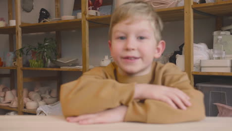 Blond-Kid-Sitting-At-A-Table-Looking-At-The-Camera-In-A-Craft-Workshop