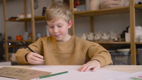 Blond-Kid-Drawing-Sitting-At-A-Table-In-A-Craft-Workshop