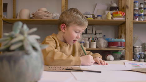 Blond-Kid-Drawing-Sitting-At-A-Table-In-A-Craft-Workshop-2