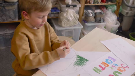 Blond-Kid-Drawing-With-Green-Pencil-Sitting-At-A-Table-In-A-Craft-Workshop-Where-Is-Signs-With-Environmental-Quotes