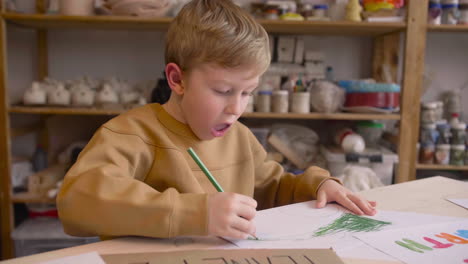 Blond-Kid-Drawing-With-Green-Pencil-Sitting-At-A-Table-In-A-Craft-Workshop-Where-Are-Signs-With-Environmental-Quotes