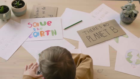 Top-View-Of-Blond-Kid-Drawing-With-Green-Pencil-Sitting-At-A-Table-In-A-Craft-Workshop-Where-Are-Signs-With-Environmental-Quotes
