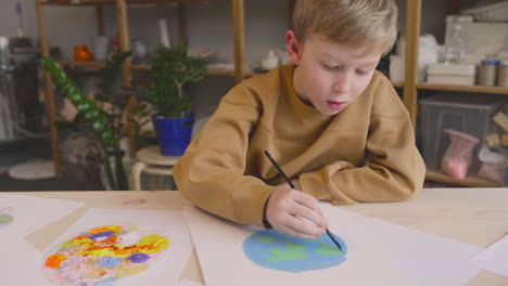Blond-Kid-Painting-A-Earth-On-A-Paper-Sitting-At-A-Table-In-A-Craft-Workshop
