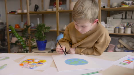 Blond-Kid-Painting-A-Earth-On-A-Paper-Sitting-At-A-Table-In-A-Craft-Workshop-Where-Are-Signs-With-Environmental-Quotes