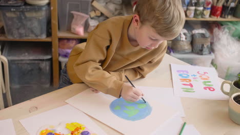 Blond-Kid-Painting-A-Earth-On-A-Paper-Sitting-At-A-Table-In-A-Craft-Workshop-Where-Are-Signs-With-Environmental-Quotes-1