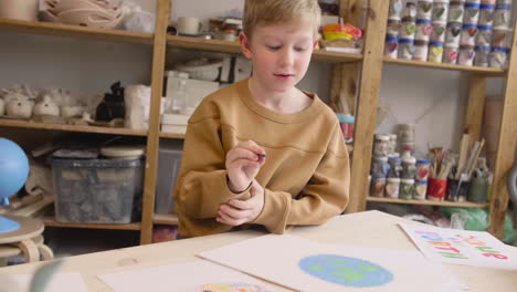 Blond-Kid-Painting-A-Earth-On-A-Paper-Sitting-At-A-Table-In-A-Craft-Workshop-Where-Are-Signs-With-Environmental-Quotes-2