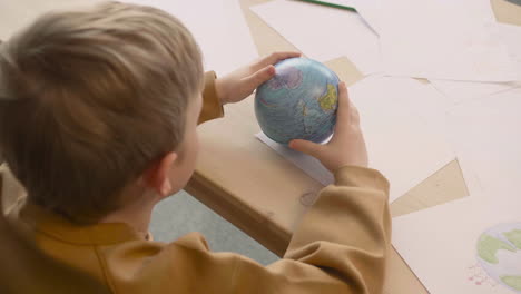 Top-View-Of-Blond-Kid-Playing-With-Globe-Sitting-At-A-Table-Where-Are-Drawings-Of-The-Earth