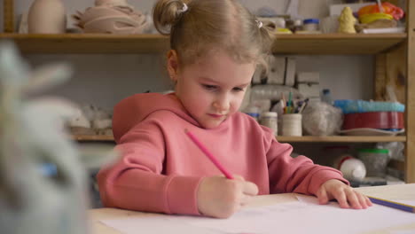 Blonde-Little-Girl-Drawing-On-A-Paper-Sitting-At-A-Table-In-A-Craft-Workshop