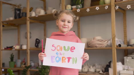 Blonde-Little-Girl-In-Craft-Workshop-Holding-A-Cardboard-Sign-With-Save-The-Earth-Phrase-1