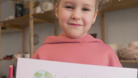 Close-Up-View-Of-A-Blonde-Little-Girl-In-Craft-Workshop-Holding-A-Cardboard-Sign-With-Save-The-Earth-Phrase