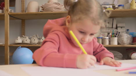Blonde-Little-Girl-Drawing-On-A-Paper-Sitting-At-A-Table-In-A-Craft-Workshop-1