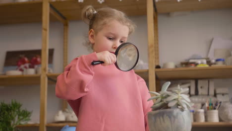 Bottom-View-Of-A-Little-Girl-Observing-A-Plant-With-A-Magnifying-Glass-Sitting-At-Table-In-A-Craft-Workshop