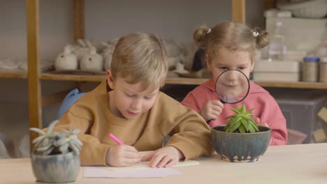 Little-Girl-Observing-A-Plant-With-A-Magnifying-Glass-Sitting-At-Table-Near-A-Friend-Who-Is-Drawing-In-A-Craft-Workshop