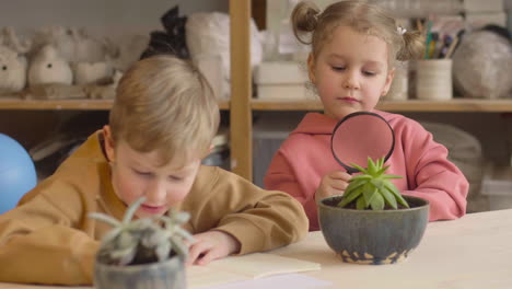 Little-Girl-Observing-A-Plant-With-A-Magnifying-Glass-Sitting-At-Table-Near-A-Friend-Who-Is-Drawing-In-A-Craft-Workshop-1