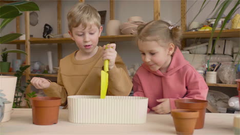 Little-Blonde-Girl-And-Blond-Kid-Preparing-The-Soil-In-A-Pot-Sitting-At-A-Table-Where-Is-Plants-In-A-Craft-Workshop