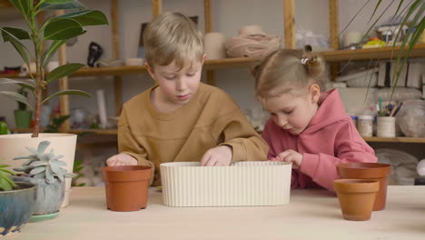 Little-Blonde-Girl-And-Blond-Kid-Preparing-The-Soil-In-A-Pot-Sitting-At-A-Table-Where-Is-Plants-In-A-Craft-Workshop-1