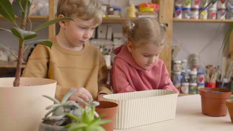 Little-Blonde-Girl-And-Blond-Kid-Preparing-The-Soil-In-A-Pot-Sitting-At-A-Table-Where-Is-Plants-In-A-Craft-Workshop-3