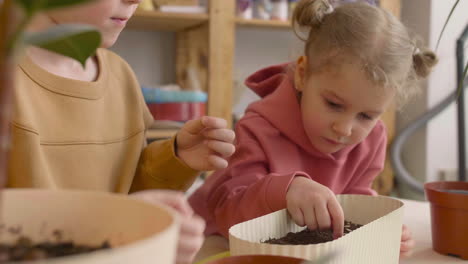 Close-Up-View-Of-Little-Blonde-Girl-And-Blond-Kid-Preparing-The-Soil-In-A-Pot-Sitting-At-A-Table-Where-Is-Plants-In-A-Craft-Workshop